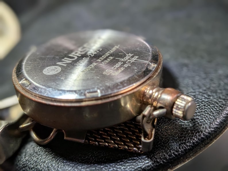 May include: A silver-toned watch case back with a metallic finish. The text reads NURSE, NMS-2475, WATER RESISTANT 10BAR, SS case back, and Made in China. A textured metal mesh strap is attached to the side, along with a small winding crown.