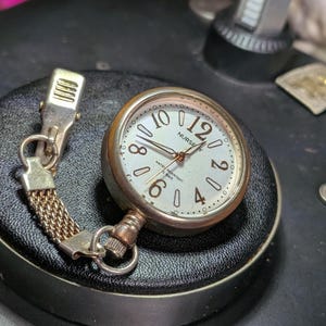 May include: A silver-toned nurse watch with a round face, featuring a white dial, black Arabic numerals for 2, 4, 6, 8, 10, and 12, and a metal clip attachment connected by a short mesh chain. The watch face displays the text NURSE and WATER RESISTANT.