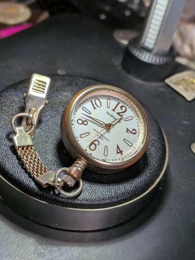 May include: A silver-toned nurse watch with a round face, featuring a white dial, black Arabic numerals for 2, 4, 6, 8, 10, and 12, and a metal clip attachment connected by a short mesh chain. The watch face displays the text NURSE and WATER RESISTANT.