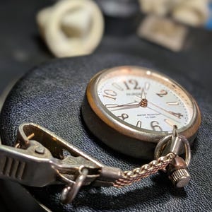 May include: A silver-toned nurse watch with a white dial, featuring large Arabic numerals at the 12, 2, 4, 6, 8, and 10 positions. The watch includes a metal clip attachment connected by a short, fine-linked chain, and the dial is marked with the text NURSE and WATER RESISTANT 10 BAR.