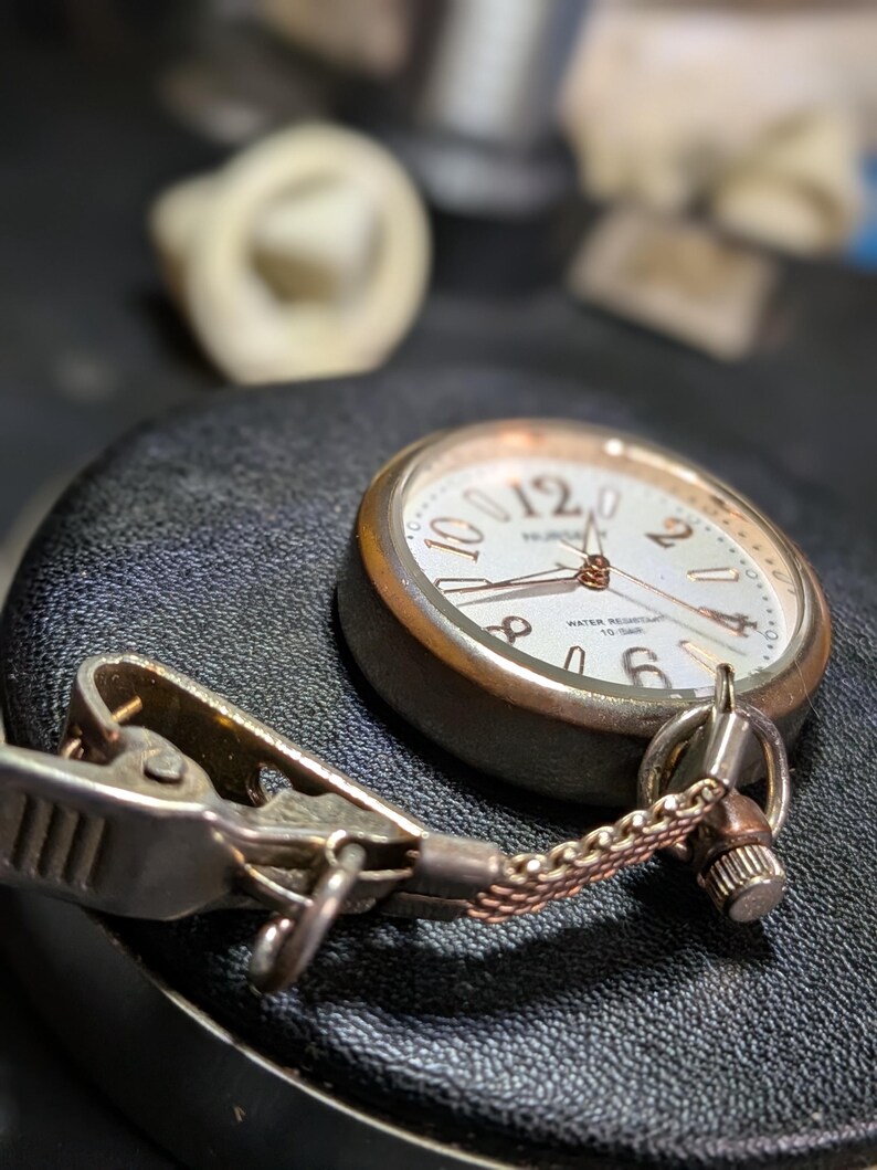 May include: A silver-toned nurse watch with a white dial, featuring large Arabic numerals at the 12, 2, 4, 6, 8, and 10 positions. The watch includes a metal clip attachment connected by a short, fine-linked chain, and the dial is marked with the text NURSE and WATER RESISTANT 10 BAR.
