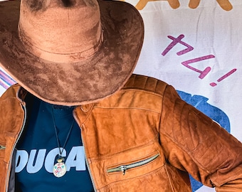 May include: A person wearing a brown leather jacket with a zippered pocket and a black t-shirt with the word "DUCATI" printed on it. The person is also wearing a brown cowboy hat.