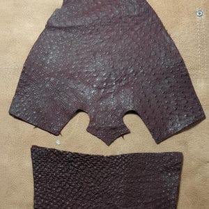 May include: Two pieces of burgundy-colored leather with a textured surface. One piece is a triangular shape with cutouts, and the other is a rectangular shape with a curved bottom edge. The leather has a bumpy texture.