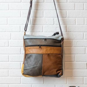 May include: A multi-colored leather crossbody bag with a long strap hangs against a white brick wall. The bag features a patchwork design in shades of brown, gray, and yellow. Sunglasses, a book, and a metal mug sit on a wooden surface.