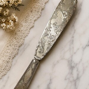 May include: An antique silver butter knife with a serrated edge and ornate floral and scrollwork engravings. The handle has a detailed design. The knife is displayed on a marble surface.