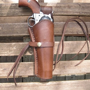 Op de afbeelding: Een bruine leren holster met een riem en een messingknop. Een zilveren revolver zit in de holster.