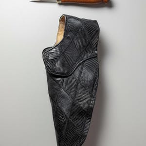 May include: A black leather holster with a diamond pattern, paired with a wood-handled metal tool. The holster has a curved shape and a textured surface. The tool is a flathead screwdriver.
