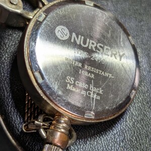 May include: The metallic back of a pocket watch featuring the engraved text Nursery, NBS-2475, Water Resistant 10BAR, SS case back, and Made in China. The circular silver-toned casing shows surface scratches and includes a metal attachment loop.