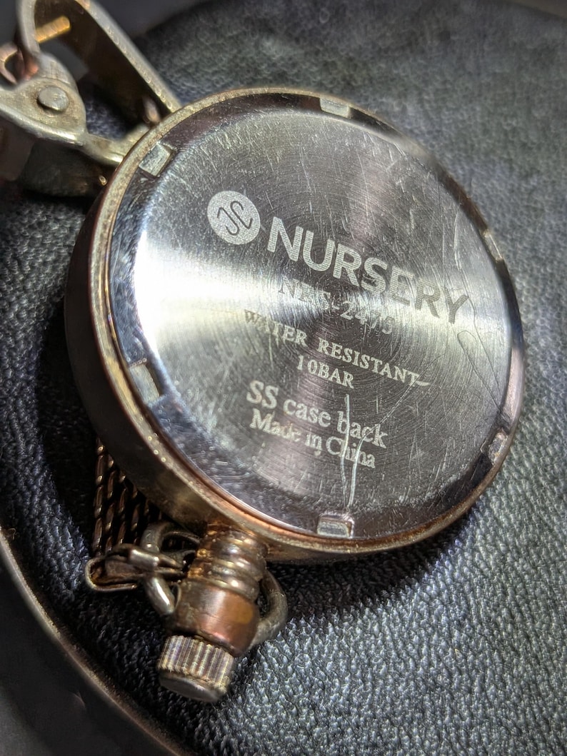 May include: The metallic back of a pocket watch featuring the engraved text Nursery, NBS-2475, Water Resistant 10BAR, SS case back, and Made in China. The circular silver-toned casing shows surface scratches and includes a metal attachment loop.
