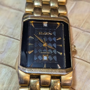 May include: Gold-toned Elgin watch with a rectangular black face. The watch face features a diamond accent and a date display. The watch band is also gold-toned. The watch face has the word "ELGIN" and "DIAMOND" printed on it.