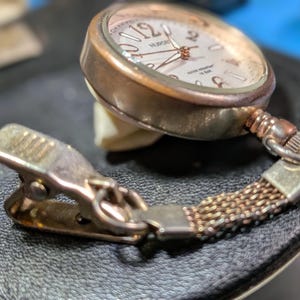 May include: A rose gold-toned nurse watch featuring a round face with large numerals, a white dial, and thin hands. The watch is attached to a short, flexible mesh chain connected to a metal spring-loaded clip for secure attachment to clothing.