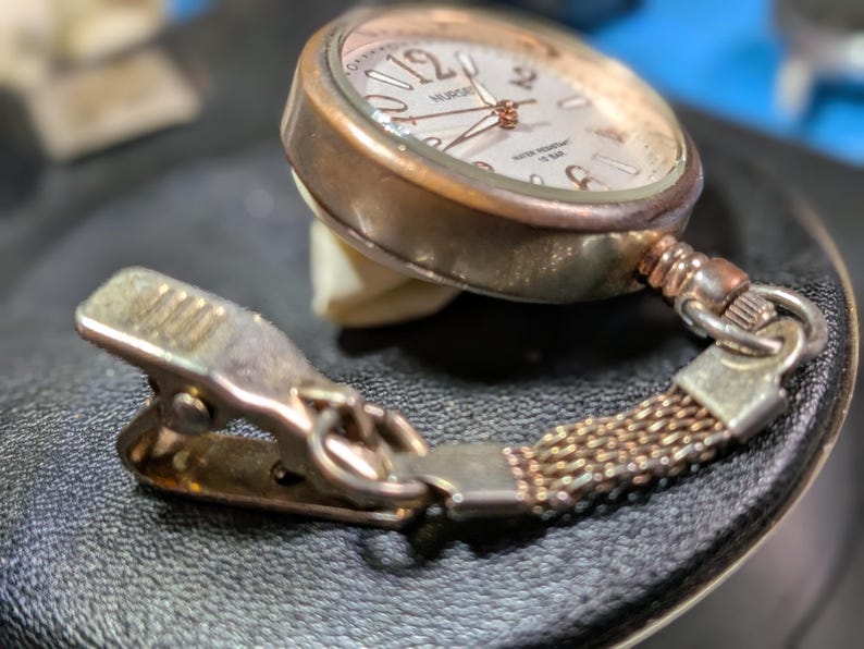 May include: A rose gold-toned nurse watch featuring a round face with large numerals, a white dial, and thin hands. The watch is attached to a short, flexible mesh chain connected to a metal spring-loaded clip for secure attachment to clothing.