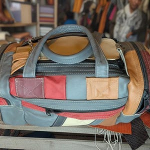 May include: A multi-colored leather duffel bag with a patchwork design. The bag features a variety of colors including red, gray, tan, and brown. It has a top handle and a zipper closure. The bag is sitting on a wooden crate.