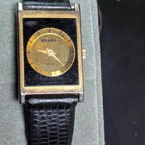 May include: A vintage Gruen wristwatch with a rectangular gold-toned case and a black face. The watch features a gold-toned circular dial with hands and the brand name. It has a black leather band.