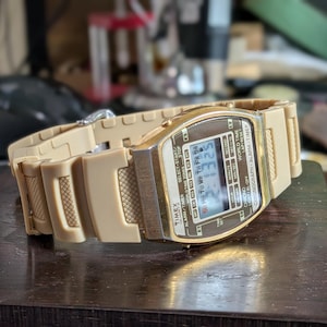 May include: A tan digital wristwatch with a metallic gold-tone bezel and a textured resin strap. The rectangular display shows the time 2:13:25, the date, and text including Timex, World Time, Chronograph, Alarm, and Timer in a black frame.