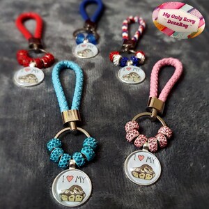 Op de afbeelding: Vijf sleutelhangers met verschillende gekleurde koorden en bedels. Elke bedel heeft een slang met de woorden "I love my" op de bedel. De bedels zijn bevestigd aan een ring met kralen. De sleutelhangers liggen op een grijs oppervlak.