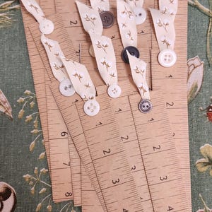 May include: Wooden rulers with inch markings, adorned with cream-colored ribbons featuring a floral pattern and various buttons. The rulers are arranged on a green floral fabric background, creating a craft or sewing theme.