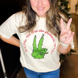 Pode incluir: Camiseta creme com o texto "It's Me, Hi, I'm the problem, it's me." em vermelho, circundando um gesto de m&atilde;o verde. A pessoa est&aacute; vestindo a camiseta e sorrindo, fazendo o sinal de paz.