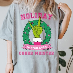 May include: A gray t-shirt with a green Grinch hand making the rock and roll sign in a pink Christmas wreath. The text "HOLIDAY est. boujee at birth CHEER MEISTER" is printed on the shirt.