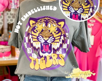 May include: Gray t-shirt with a purple and yellow tiger graphic. The tiger has gold glitter eyes and the text "Not Embellished" is written above the tiger. The text "Tigers" is written in a retro font below the tiger. The text "The Yellow Daffodil Boutique" is written in the bottom right corner of the shirt.