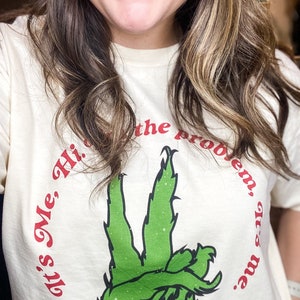 Pode incluir: Camiseta creme com um gesto de m&atilde;o verde com um design semelhante ao Grinch. O texto "It's Me, Hi, I'm the problem, it's me." &eacute; impresso em vermelho ao redor da m&atilde;o. Uma camiseta casual e divertida.