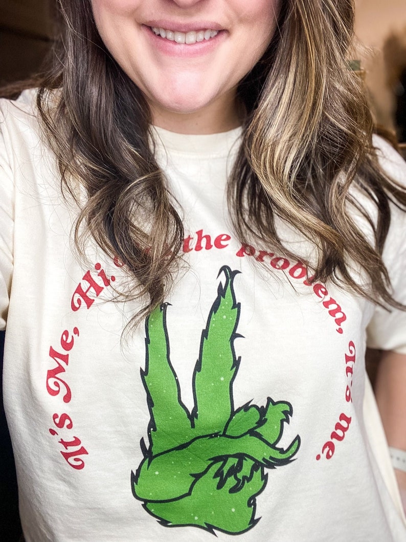 Pode incluir: Camiseta creme com um gesto de m&atilde;o verde com um design semelhante ao Grinch. O texto "It's Me, Hi, I'm the problem, it's me." &eacute; impresso em vermelho ao redor da m&atilde;o. Uma camiseta casual e divertida.
