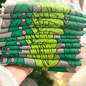 May include: A stack of gray t-shirts with a green and pink Christmas tree design. The text "HOLLIDAY" is visible on the top shirt.