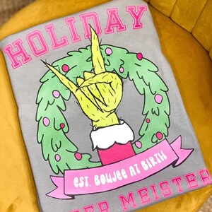 May include: Gray t-shirt with a pink "HOLIDAY" graphic. The graphic features a green wreath with a yellow hand giving the peace sign. The text below the wreath reads "est. boujee at birth" and "CHEER MEISTER".