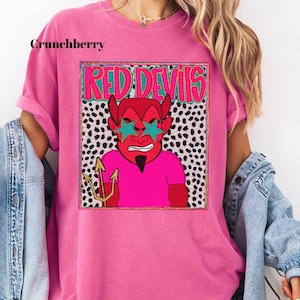 Puede incluir: Camiseta rosa con un gráfico de un diablo rojo con gafas de sol en forma de estrella y un tridente. El texto "RED DEVILS" está encima de la ilustración del diablo. La marca "Crunchberry" está impresa en la parte superior izquierda.
