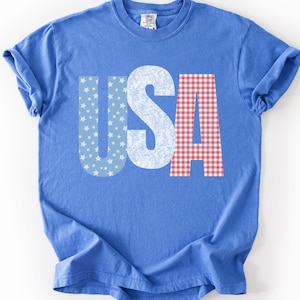 Puede incluir: Una camiseta azul con la palabra "USA" en blanco. La letra "U" está llena de estrellas blancas sobre un fondo azul, la "S" tiene un estampado floral blanco y la "A" tiene un estampado de cuadros rojos y blancos.