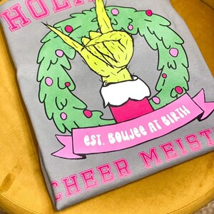May include: Gray t-shirt with a pink "HOLIDAY" graphic. The graphic features a green hand making a rock and roll gesture inside a green wreath with pink ornaments. The text "est. boujee at birth" is on a pink banner below the wreath. The text "CHEER MEIST" is printed in pink at the bottom of the shirt.