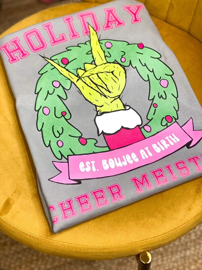 May include: Gray t-shirt with a pink "HOLIDAY" graphic. The graphic features a green hand making a rock and roll gesture inside a green wreath with pink ornaments. The text "est. boujee at birth" is on a pink banner below the wreath. The text "CHEER MEIST" is printed in pink at the bottom of the shirt.
