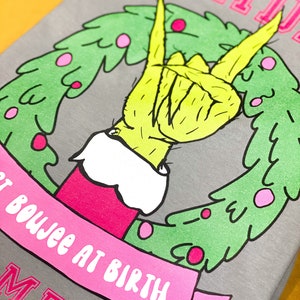 May include: A gray t-shirt with a pink and green Christmas wreath design. The wreath features a green Grinch hand giving the peace sign. The text "HOLIDAY" is at the top of the shirt and "NOT BOUJEE AT BIRTH" is on a pink ribbon below the wreath. The text "MEIS" is at the bottom of the shirt.