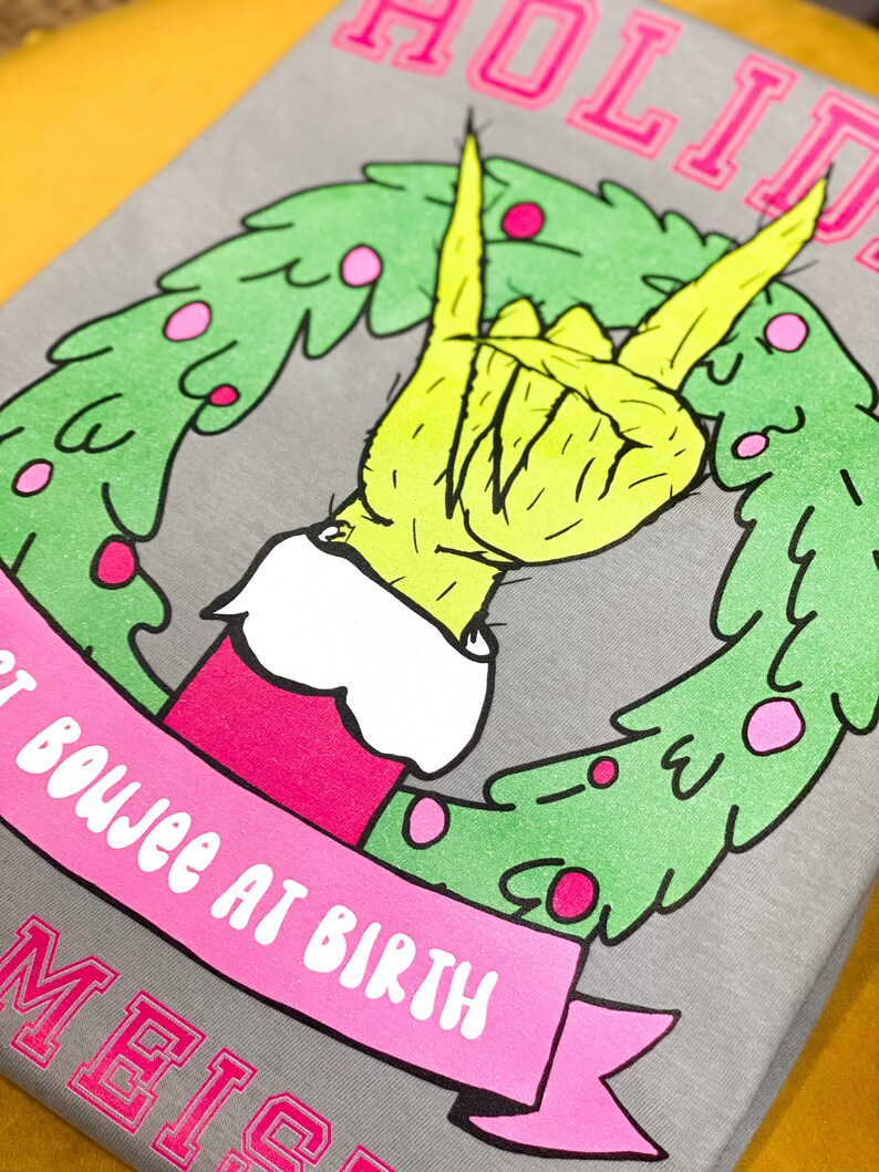 May include: A gray t-shirt with a pink and green Christmas wreath design. The wreath features a green Grinch hand giving the peace sign. The text "HOLIDAY" is at the top of the shirt and "NOT BOUJEE AT BIRTH" is on a pink ribbon below the wreath. The text "MEIS" is at the bottom of the shirt.