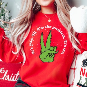 Pode incluir: Uma camisola vermelha de manga comprida com o texto "It's Me, Hi, I'm the problem, It's me." num design circular. O design apresenta uma m&atilde;o verde a fazer o sinal de paz. Um artigo festivo de f&eacute;rias.
