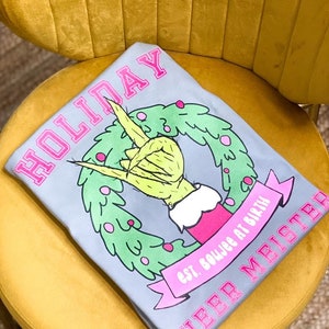 May include: Gray t-shirt with a pink "HOLIDAY" graphic. The graphic features a green hand making a rock and roll sign inside a green wreath. The text "est. boujee at birth" and "CHEER MISTER" are also printed in pink.