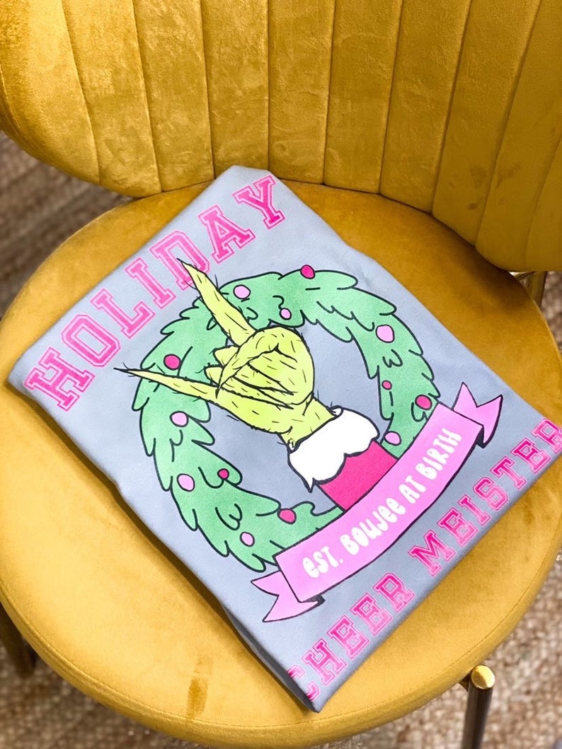 May include: Gray t-shirt with a pink "HOLIDAY" graphic. The graphic features a green hand making a rock and roll sign inside a green wreath. The text "est. boujee at birth" and "CHEER MISTER" are also printed in pink.