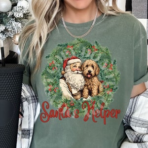 May include: A green t-shirt with a vintage illustration of Santa Claus and a dog in a wreath. The text "Santa's Helper" is printed in red below the image.