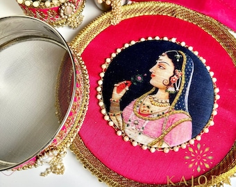 May include: A collection of decorative Indian items, including a pink and gold tray with a woman's portrait, a stainless steel sieve, and two stainless steel cups. The items are adorned with pearls and gold trim.