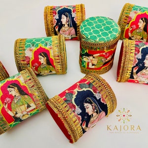May include: Six decorative cylindrical boxes with gold trim and colorful fabric featuring images of women in traditional Indian clothing. The boxes are decorated with floral patterns and have a green, red, and blue color scheme. The text "Kajora The House of Happiness" is visible at the bottom of the image.