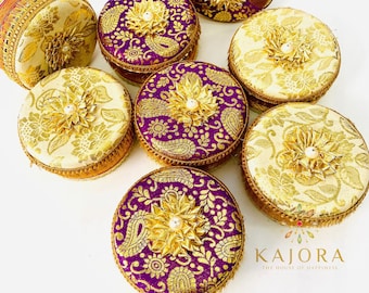 May include: An assortment of round, decorative boxes with ornate gold and purple fabric. Each box features a gold floral embellishment with a pearl center. The boxes are arranged on a white surface, showcasing their intricate designs and textures.