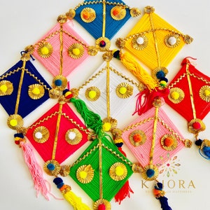 May include: A set of nine colorful diamond-shaped wall hangings made of yarn and decorated with gold accents. Each hanging features a tassel and pom-poms. The hangings are arranged in a staggered pattern on a white background. The text "KAHORA THE HOUSE OF HAPPINESS" is visible in the bottom right corner of the image.