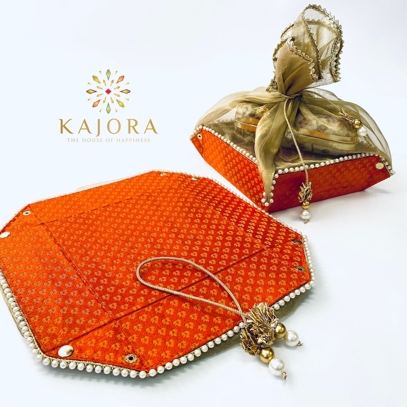 Pu&ograve; includere: Un set di due scatole regalo in tessuto arancione con bordino di perle e nappina dorata. Le scatole sono piegate a piatto e hanno un motivo decorativo. Il testo "Kajora The House of Happiness" &egrave; visibile nell'angolo in alto a sinistra dell'immagine.