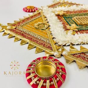 May include: A decorative wall hanging with a geometric design in shades of red, gold, and green. The hanging is adorned with white fabric flowers and pearl accents. Two small, red, round candle holders with gold accents are also included.