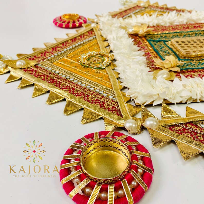 May include: A decorative wall hanging with a geometric design in shades of red, gold, and green. The hanging is adorned with white fabric flowers and pearl accents. Two small, red, round candle holders with gold accents are also included.