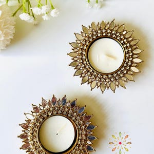 May include: Two circular tea light candle holders featuring a metallic gold frame adorned with a sunburst pattern of small mirrors and clear rhinestones. Each holder contains a white wax candle. The text KAJORA THE HOUSE OF HAPPINESS is visible.