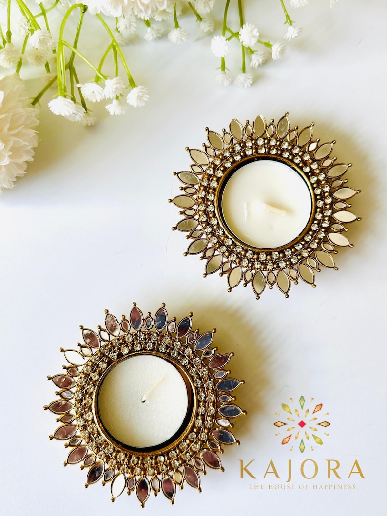 May include: Two circular tea light candle holders featuring a metallic gold frame adorned with a sunburst pattern of small mirrors and clear rhinestones. Each holder contains a white wax candle. The text KAJORA THE HOUSE OF HAPPINESS is visible.