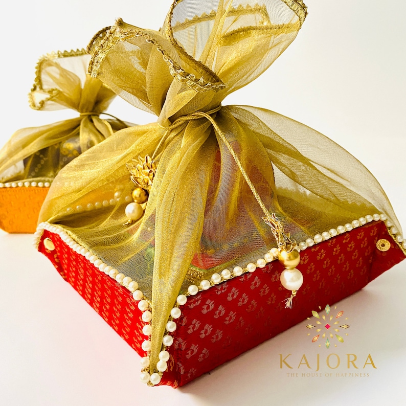 Pu&ograve; includere: Una scatola regalo rossa e dorata con un tessuto trasparente dorato legato intorno. La scatola &egrave; decorata con perle bianche e ha un piccolo fiocco dorato appeso. Il testo "Kajora The House of Happiness" &egrave; stampato nell'angolo in basso a destra dell'immagine.