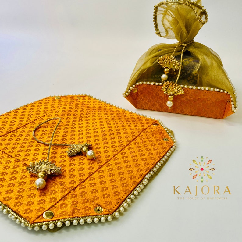 Pu&ograve; includere: Un set di due scatole decorative di colore arancione brillante con un motivo dorato. Le scatole sono rivestite di perle bianche e hanno un fiocco dorato. Le scatole sono etichettate "Kajora, The House of Happiness".