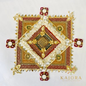 May include: A square-shaped decorative piece with intricate gold and red embroidery, surrounded by five small red candle holders. The center of the piece features a white, fluffy design with gold accents. The text "Kajara The House of Happiness" is visible at the bottom of the image.